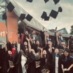 A group of CU Coventry students celebrating their graduation day by throwing their caps in the air.