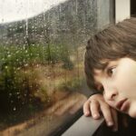 An upset young boy sitting indoors by a window, watching the rain
