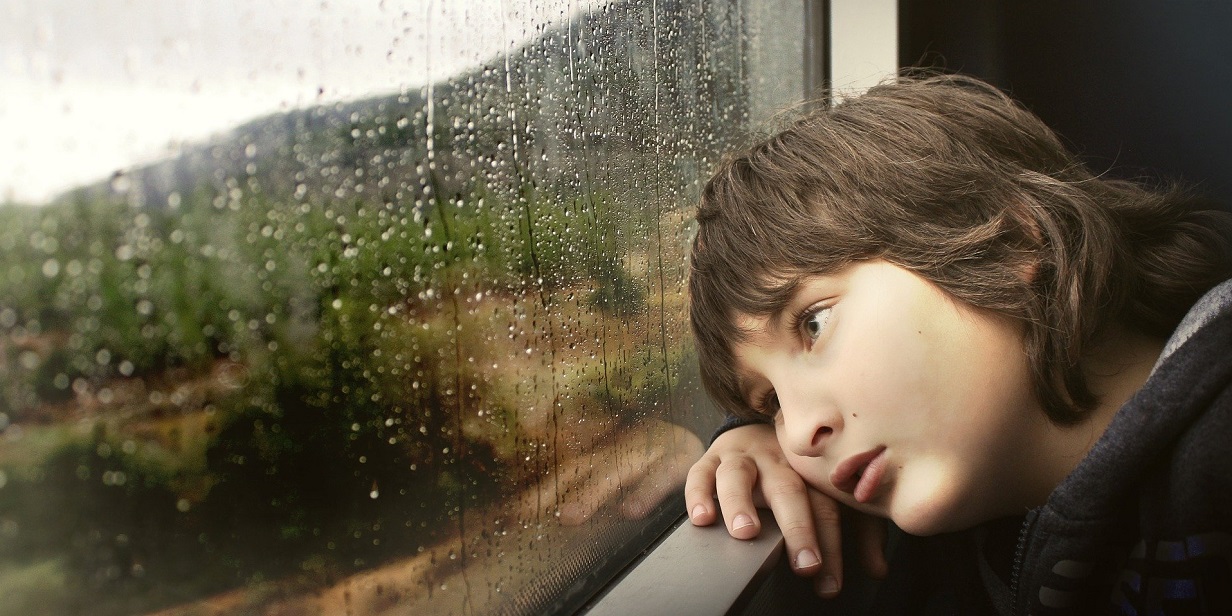 An upset young boy sitting indoors by a window, watching the rain
